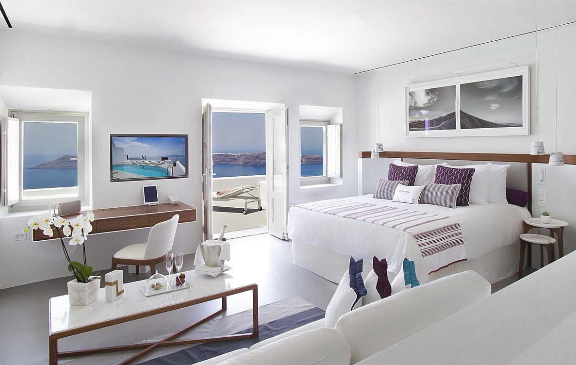 Grace Santorini room, with white washed walls, polished gray concrete floor, and white bed with purple cushions