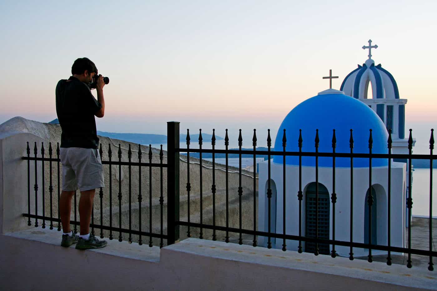 Young man photographs a blue-domed church on Santorini at sunset.