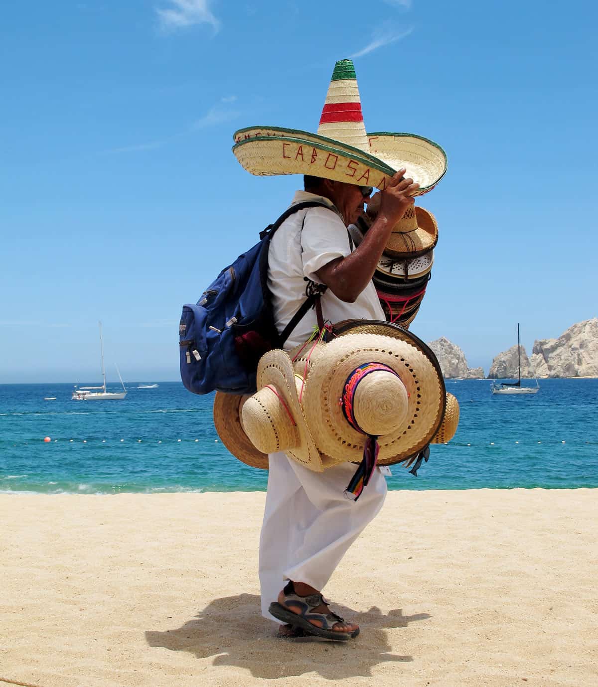 A Cabo San Lucas beach vendor, dressed in white, carries loads of hats.