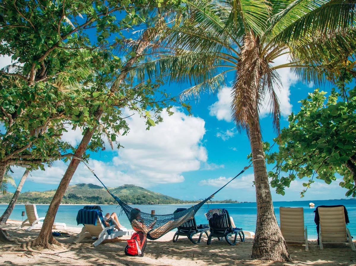 Woman swings in a hammock, reading a book, on a Fiji beach.