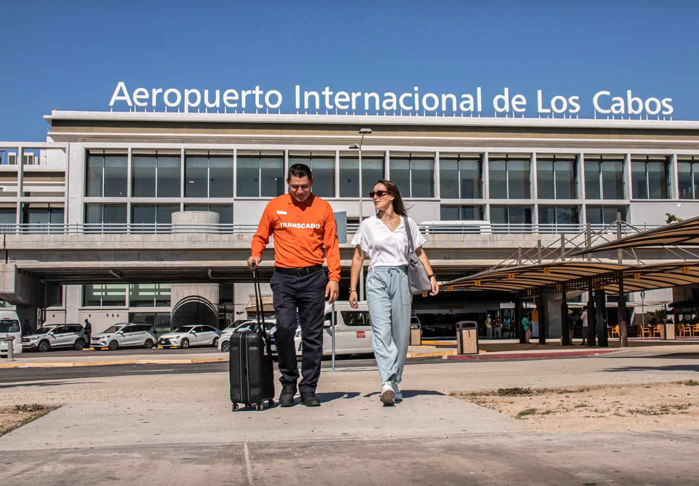 TransCabo driver in bright orange shirt assists a passenger with luggage outside the Los Cabos International Airport.
