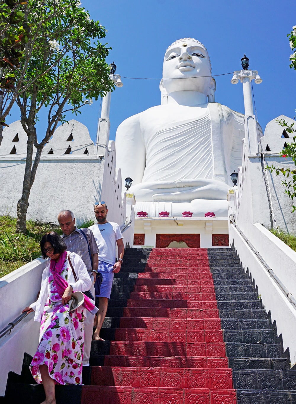 Visitors walk barefoot down red-and-black steps beneath the enormous white seated Buddha at Bahiravokanda Vihara in Kandy.