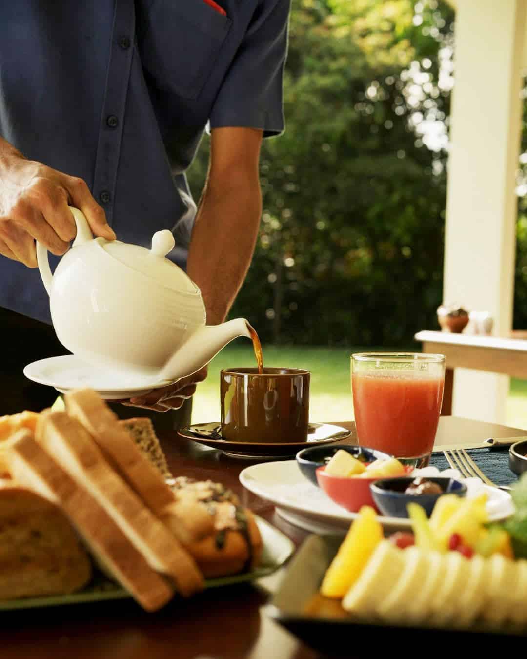Server pouring white pot of tea and breakfast spread of toast and fruit