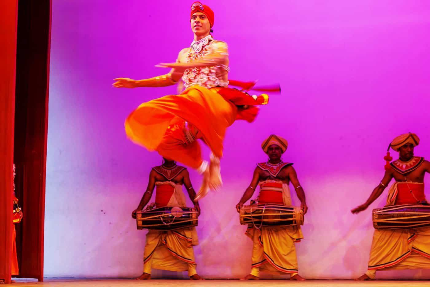 A Kandyan dancer in an orange costume leaps to the beat of pounding drums.