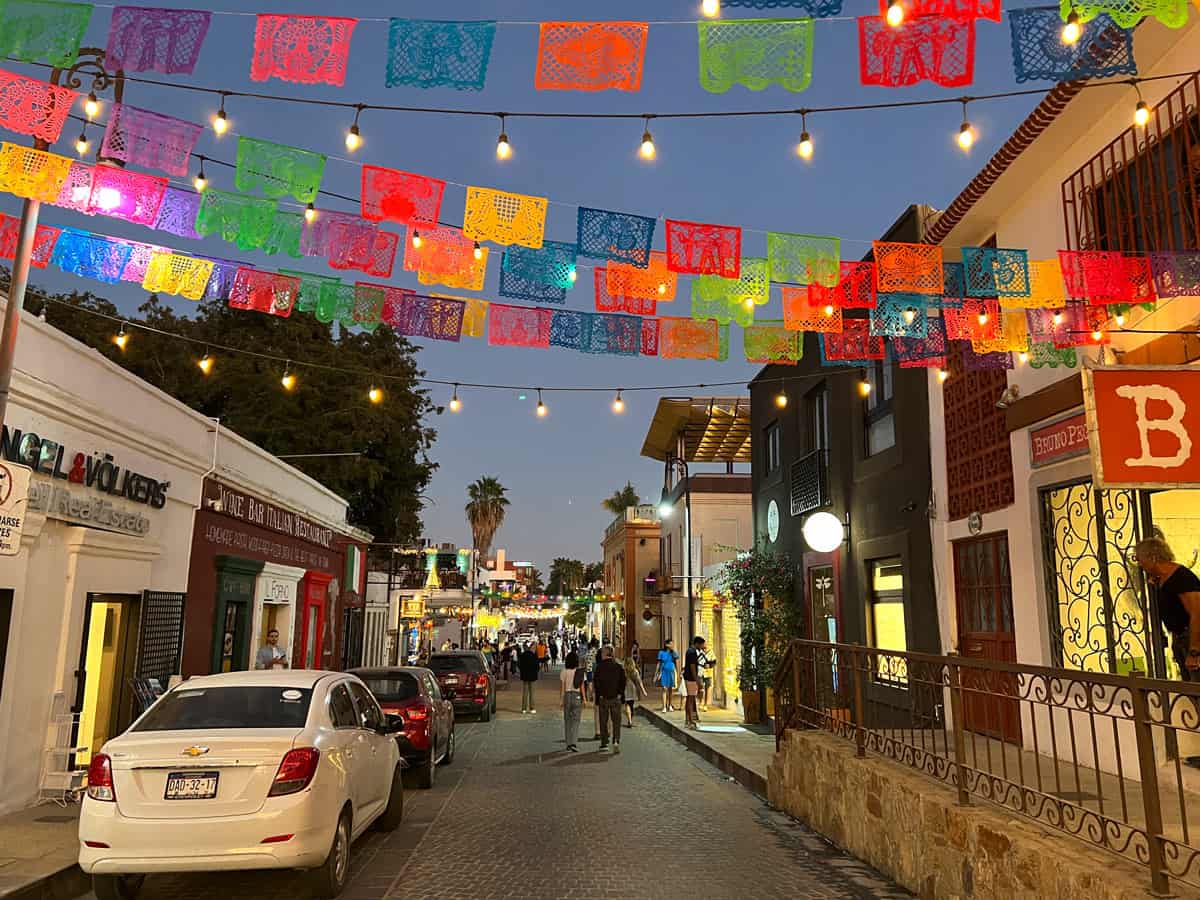Colorful papel picado banners and glowing string lights decorate a street in San Jose del Cabo's historic art district at dusk.