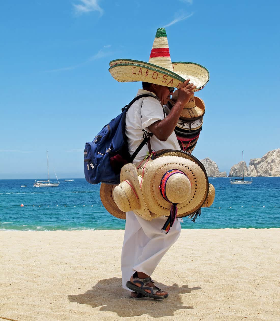A beach vendor carries a colorful collection of straw hats and sombreros along Medano Beach in Cabo San Lucas, with Land's End in the background.