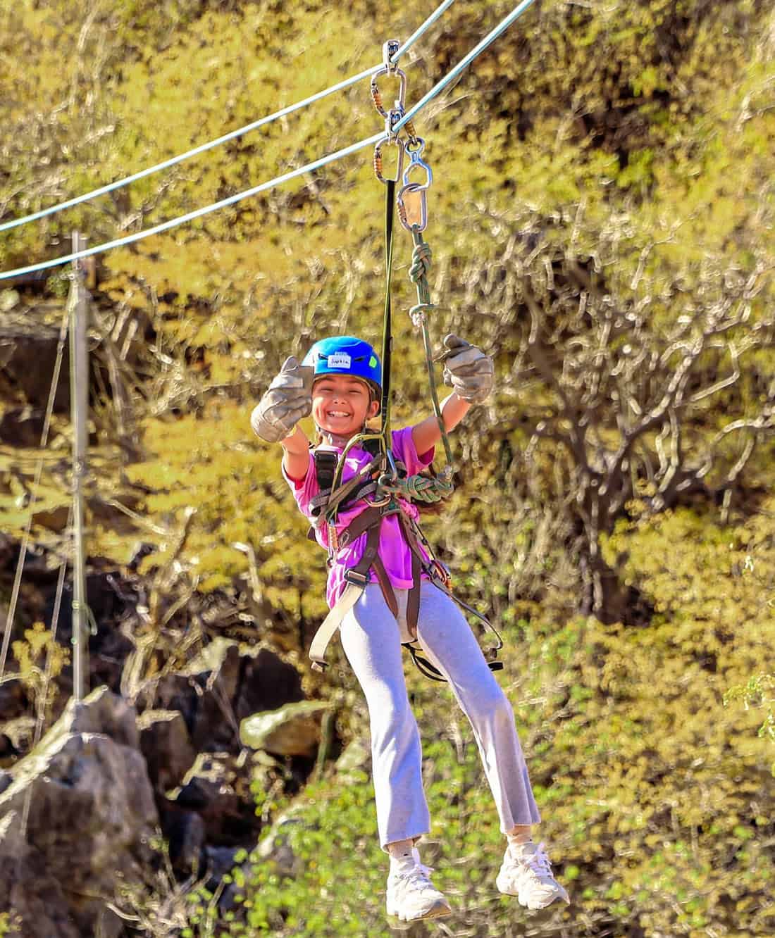 Smiling young child, wearing a blue helmet, pink T-shirt and safety harness, ziplines over a rocky canyon in Cabo.