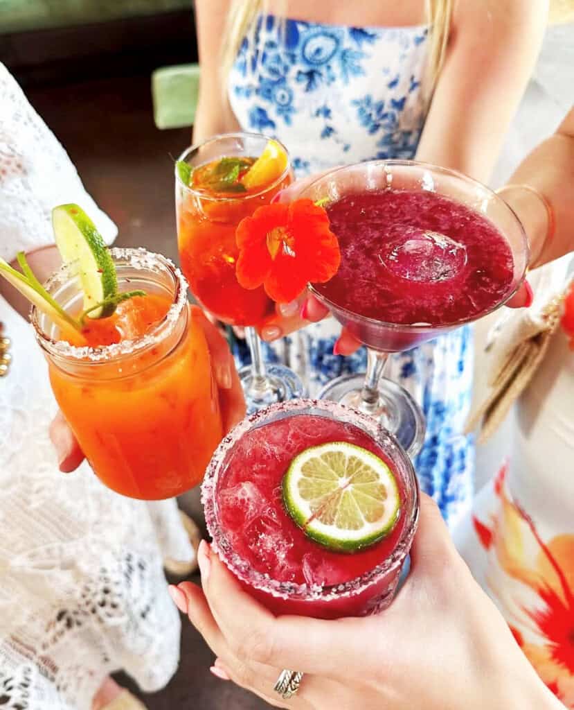 Four red and orange cocktails garnished with lime, mint and an edible flower, raised for a toast at Flora Farms in Los Cabos