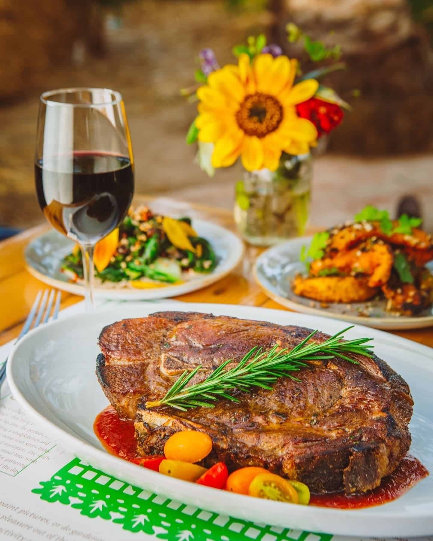 Juicy grilled bone-in steak topped with fresh rosemary, served with colorful cherry tomatoes, side dishes and red wine at Flora Farms