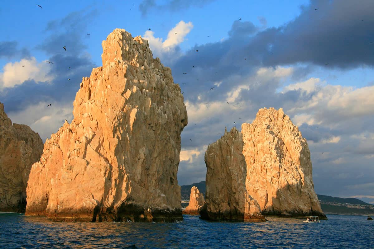 Towering golden granite rock formations, at Landโs End in Cabo San Lucas, glow in the late-afternoon sunlight beneath dramatic clouds.