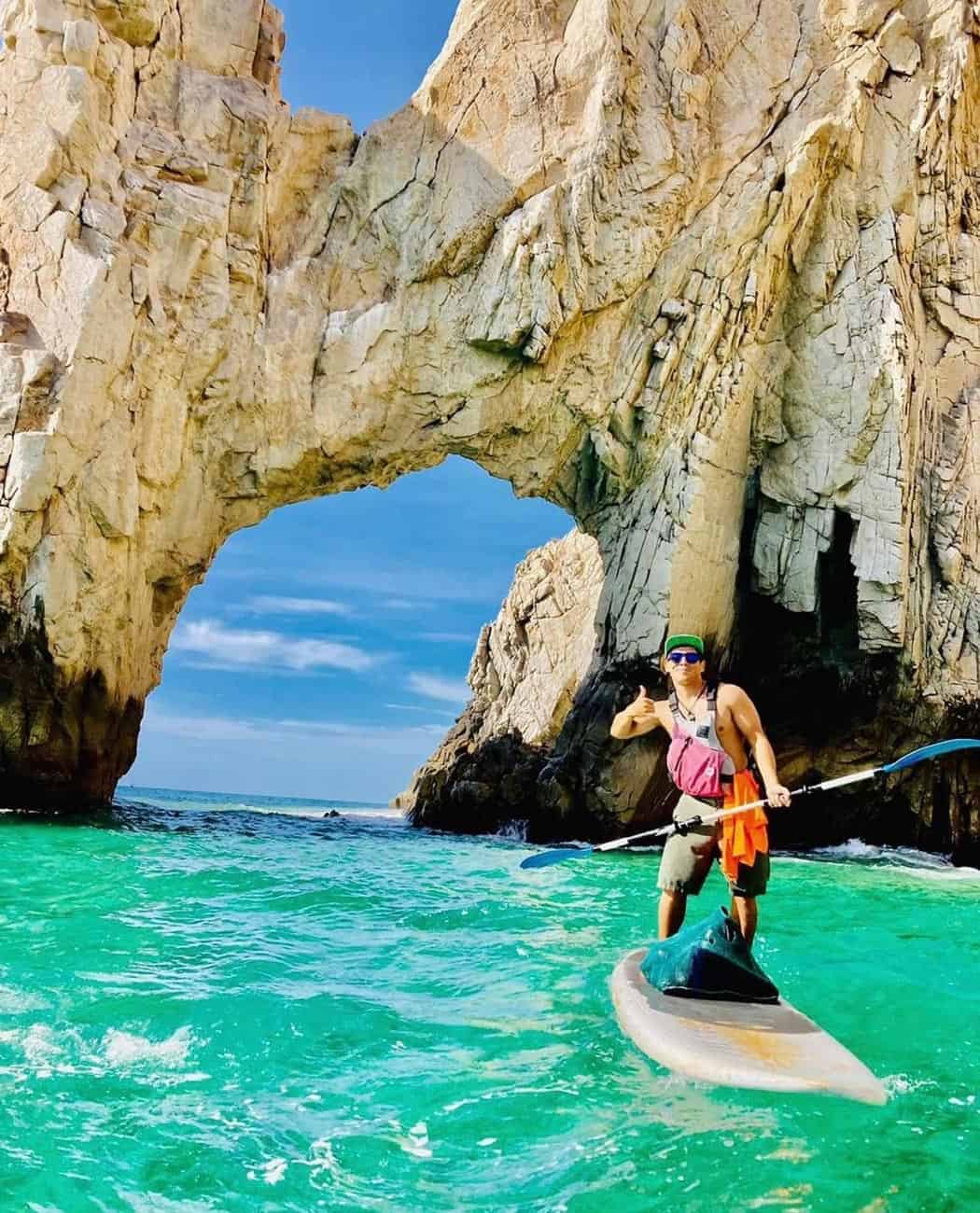 Man on a stand-up paddleboard gives a thumbs-up in front of Cabo San Lucas Arch.