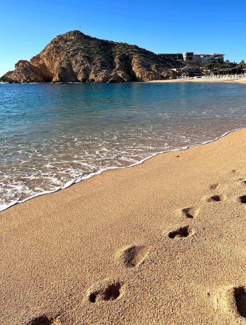 Footprints in the golden sand at Santa Maria Beach in Cabo San Lucas, with turquoise water and rugged rocky cliffs across the bay