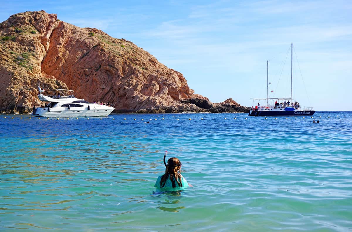 Snorkeler wading into turquoise waters at Santa Maria Bay, with a yacht, sailboat and rugged golden cliffs in the background