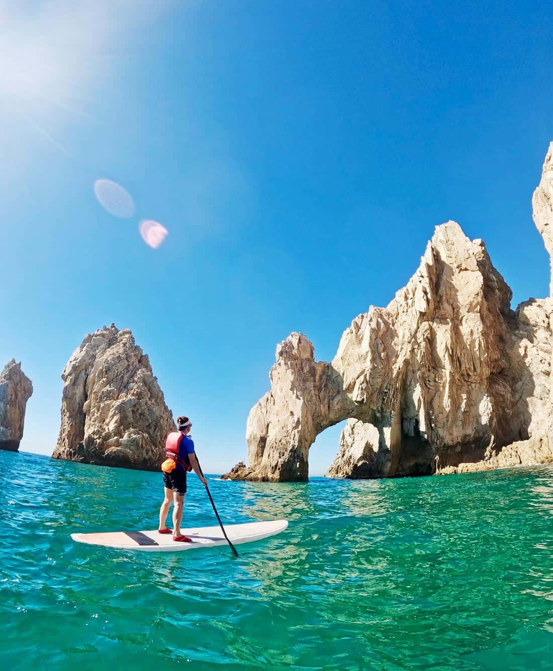 Woman paddleboards across turquoise water toward the famous Arch at Landโs End in Cabo San Lucas.
