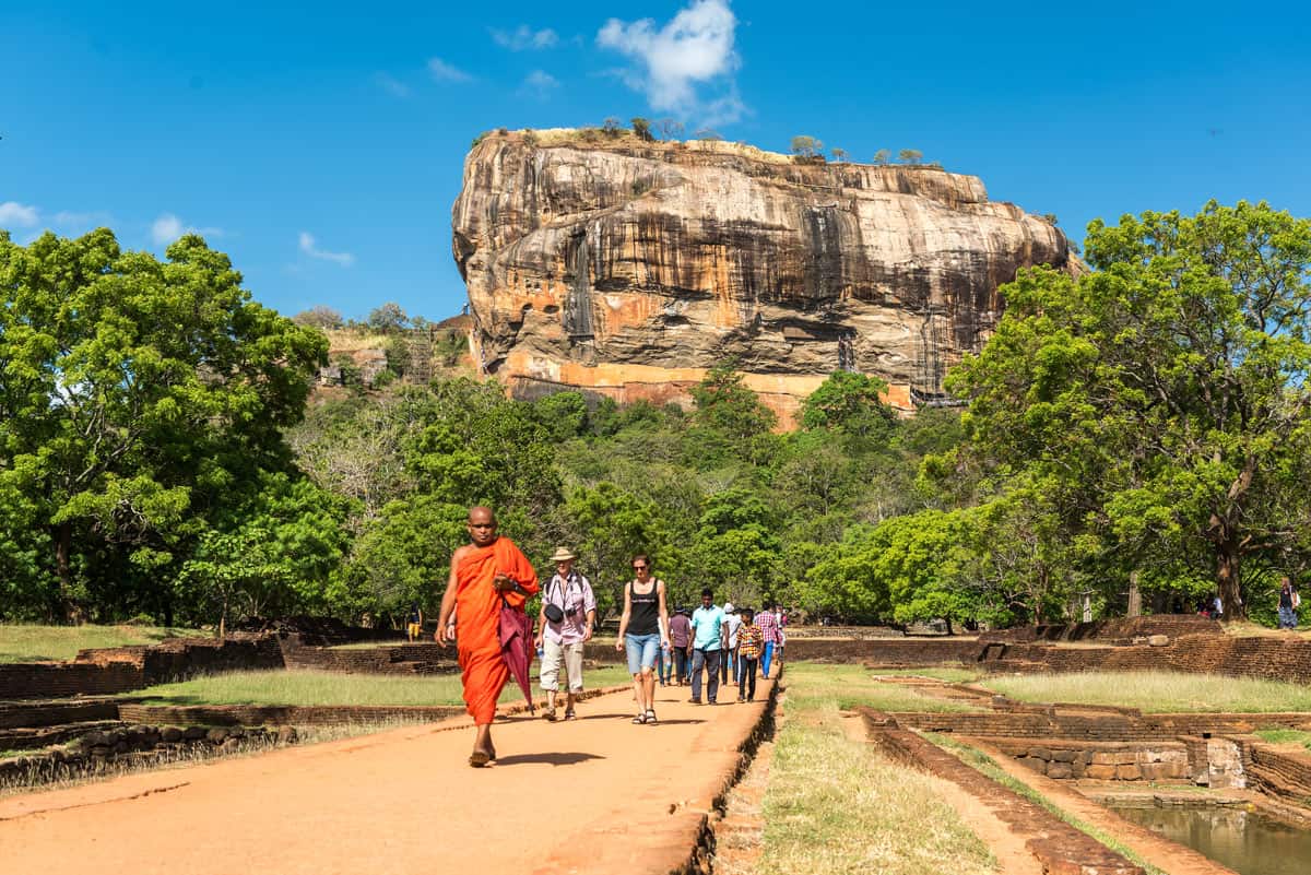 Red-robed monk and visitors walk at the base of the soaring golden rock of Sigiriya