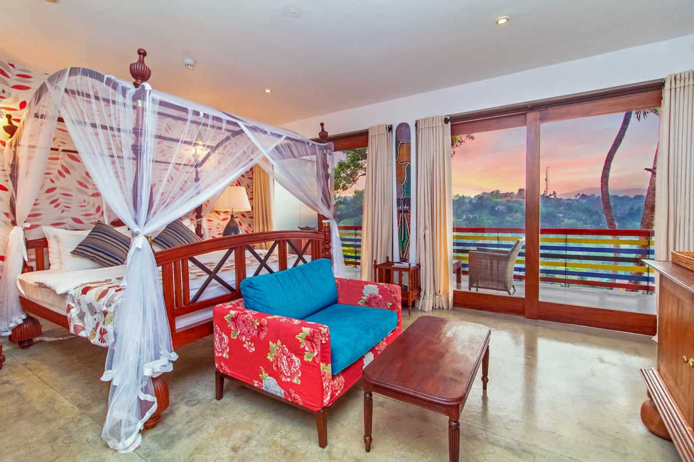 Guest room at The Elephant Stables with four poster bed, draped with white mosquito netting, and red loveseat
