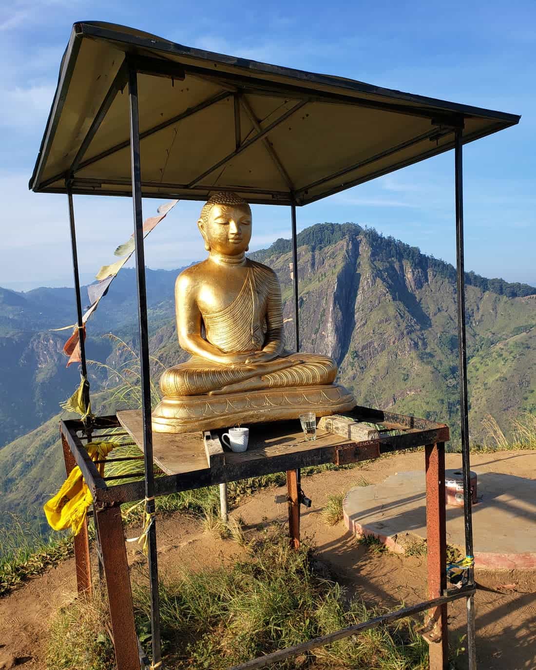 A seated gold Buddha statue sits on a platform on top of Little Adam's Peak, with green mountains rising behind it.