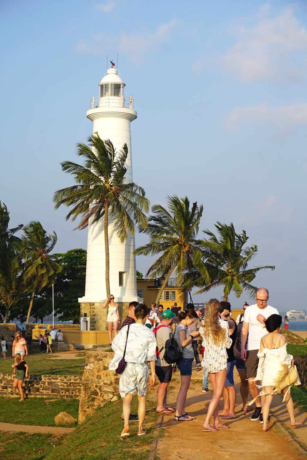 The tall white Galle Lighthouse, bathed with a golden hue at sunset, with visitors taking photos along the fort ramparts