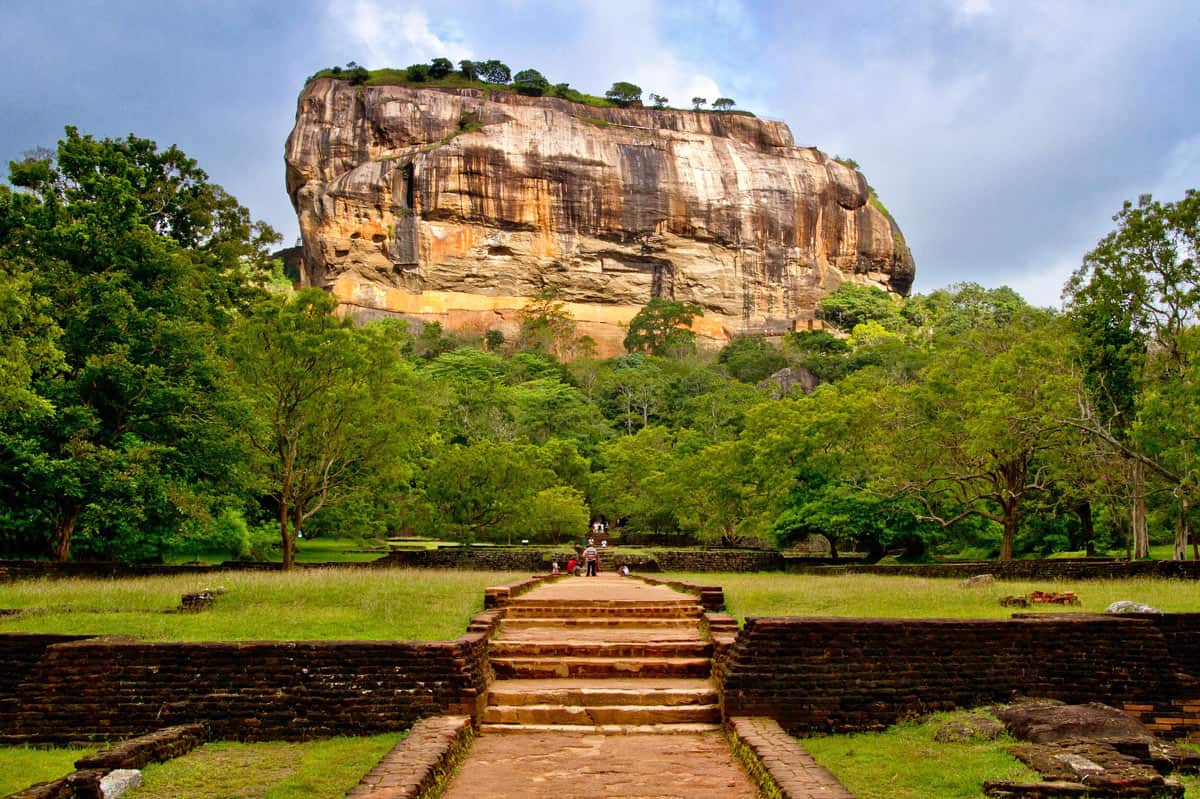 Huge gold-brown rock towering above green forest, with a dirt path leading to the rock