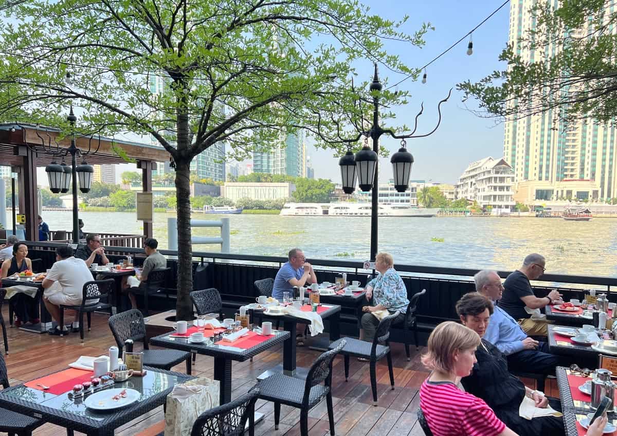 Guests sit on the outdoor breakfast terrace at NEXT2 Cafรฉ at Shangri-La Bangkok, where tables overlook boats on the Chao Phraya River.