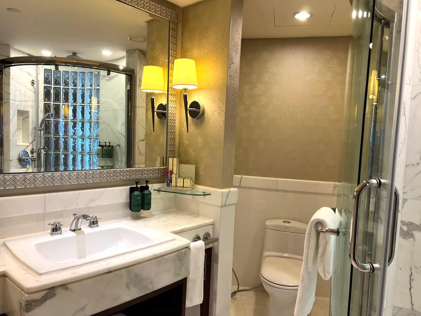White marble bathroom in a Shangri-La Bangkok Deluxe River View Room, with a large vanity, wall mirror and curved glass shower
