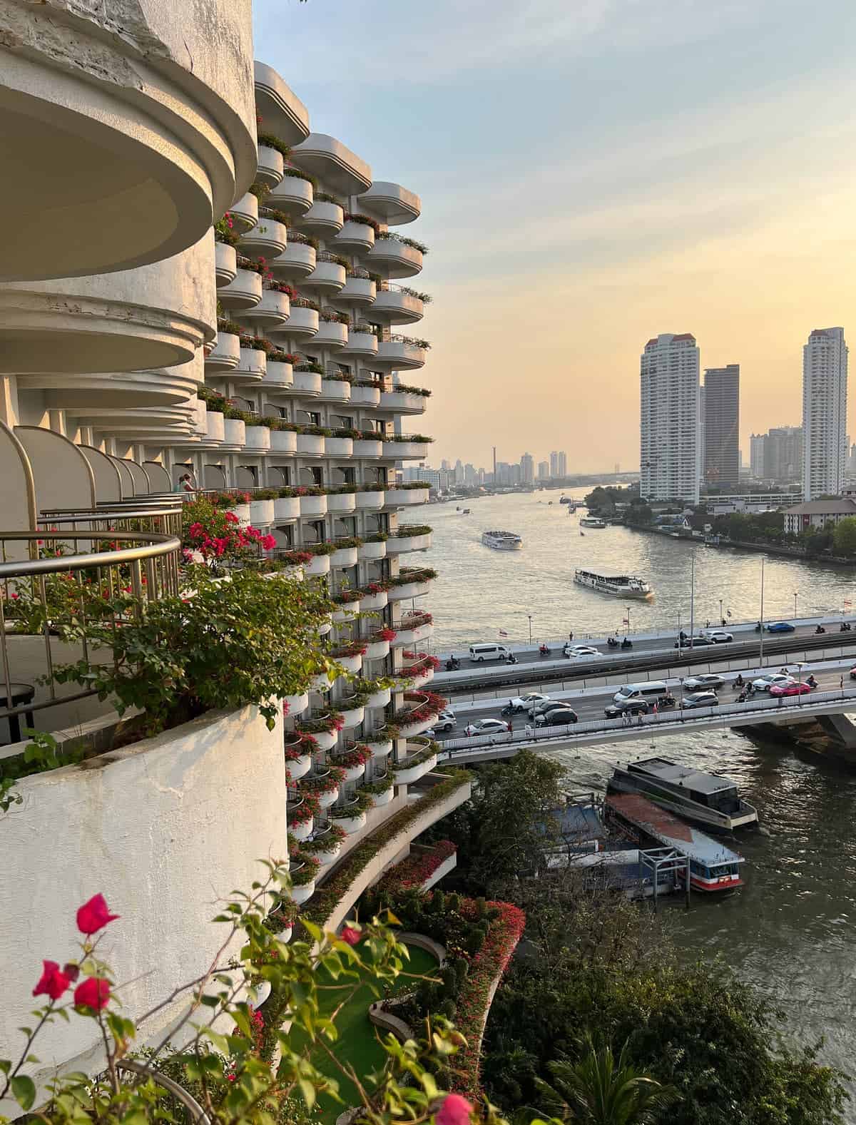View from a Krungthep Wing balcony over flower-filled curved balconies and the Chao Phraya River, with boats and Bangkok high-rises