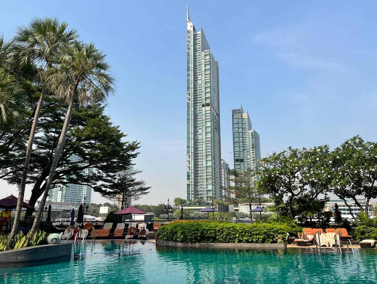 Shangri-La Bangkokโs main swimming pool surrounded by palm trees, tropical gardens and loungers, with views of Bangkok skyscrapers
