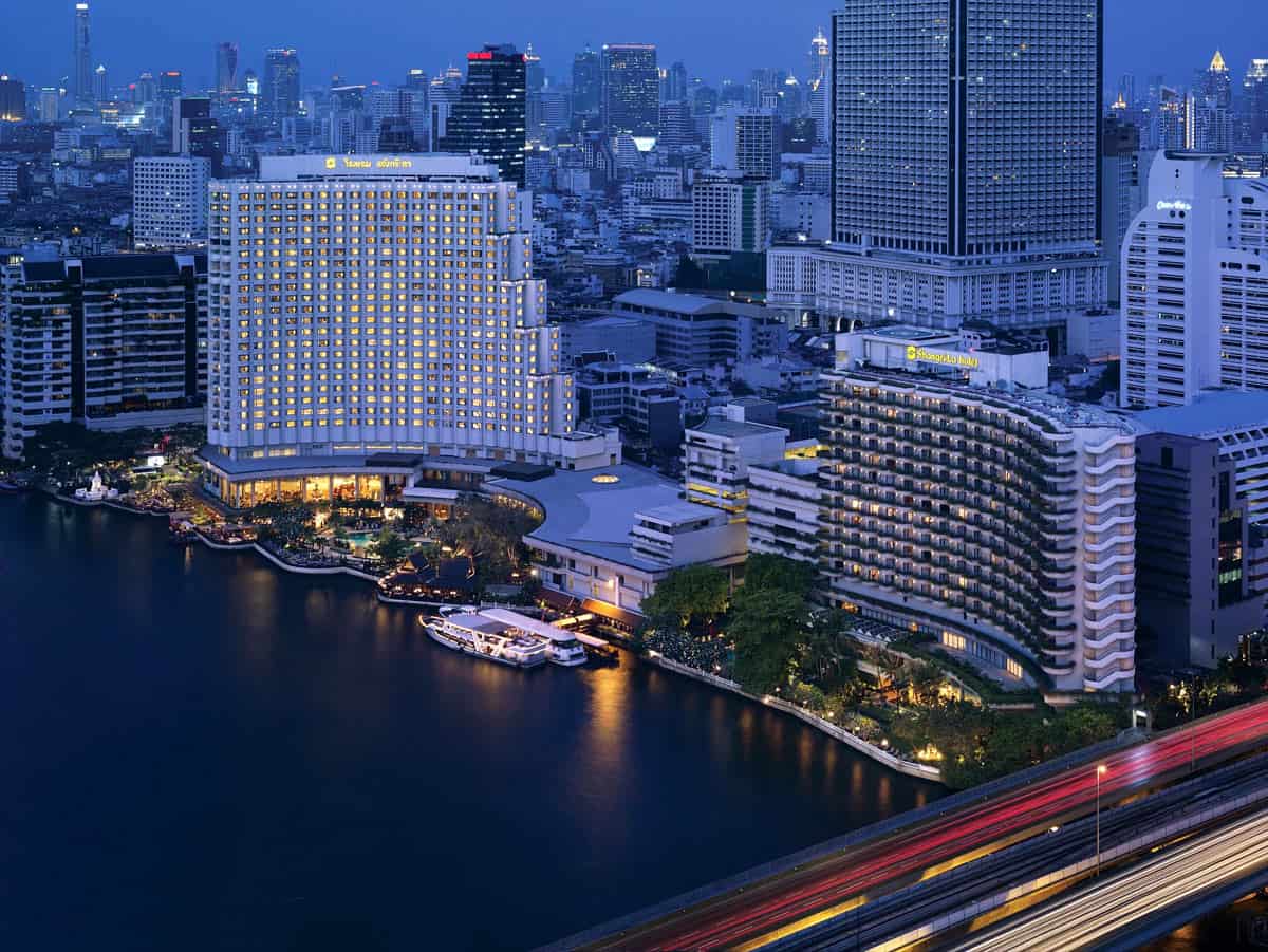 Shangri-La Bangkok illuminated at night beside the Chao Phraya River, with the Bangkok skyline and streaks of city traffic lights