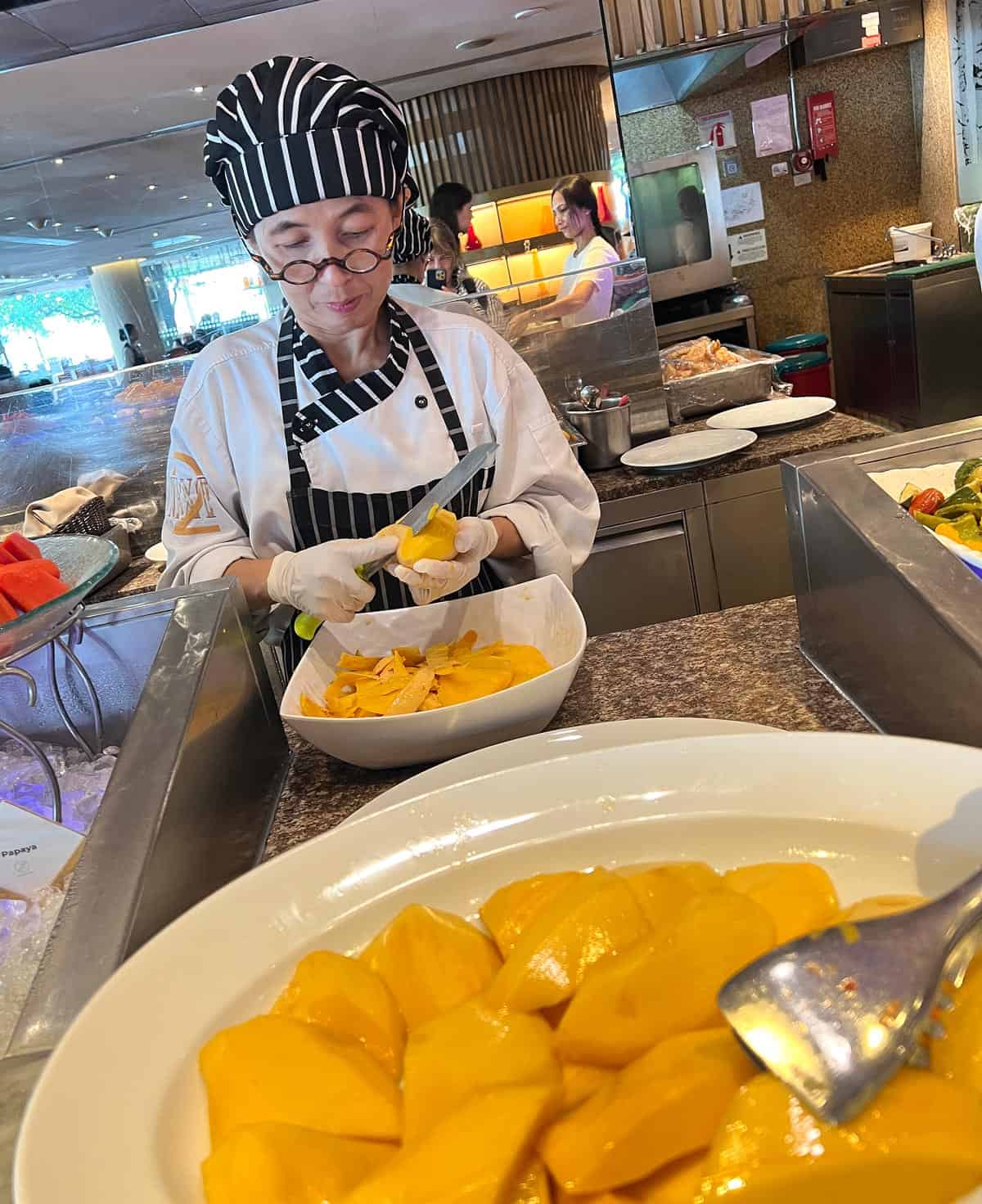 A female chef slices fresh ripe mango at the NEXT2 Cafรฉ breakfast buffet at Shangri-La Bangkok.