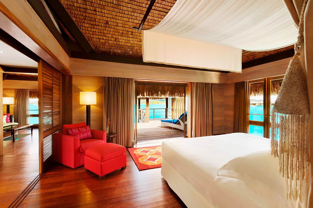 Bedroom of Overwater Bungalow at St. Regis Bora Bora, with wood floors and a red lounge chair and footstool