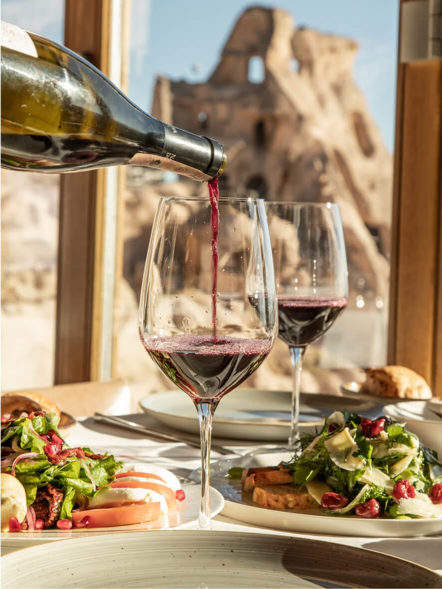 Two glasses of red wine, plates of food and a backdrop of rocky Uchisar Castle