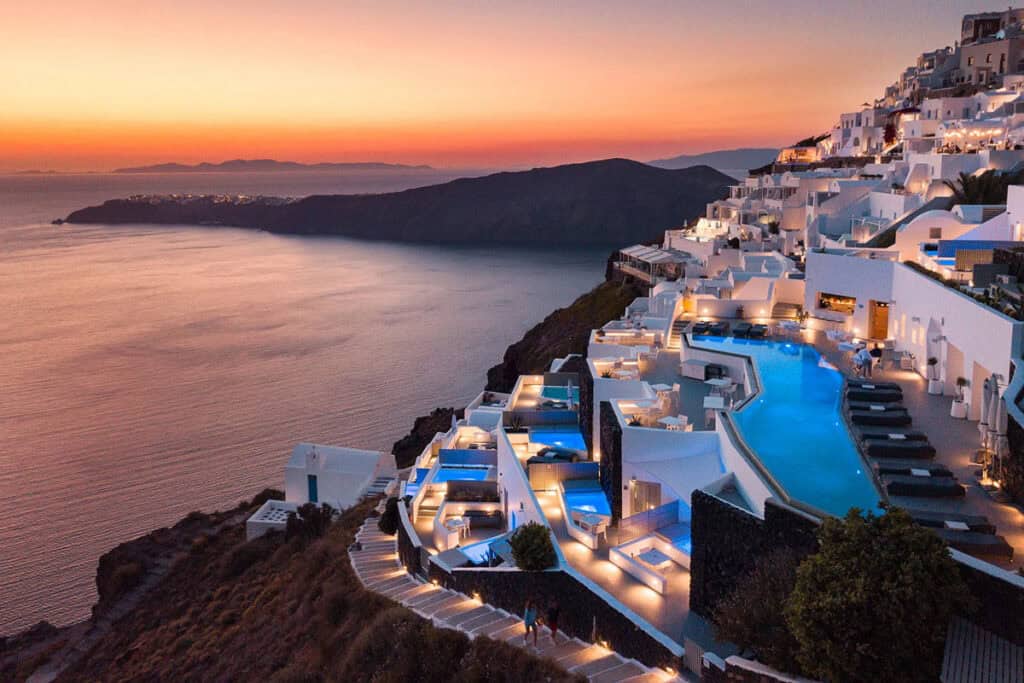 Whitewashed cliffside hotel with glowing blue pools, overlooking the sea at sunset, beneath an orange sky