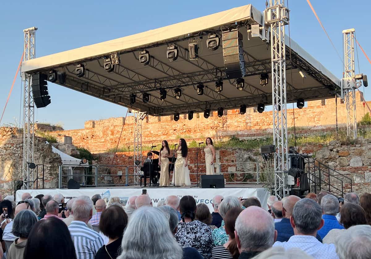 Three sopranos in gold evening dresses perform on stage in an old fortress at sunset.