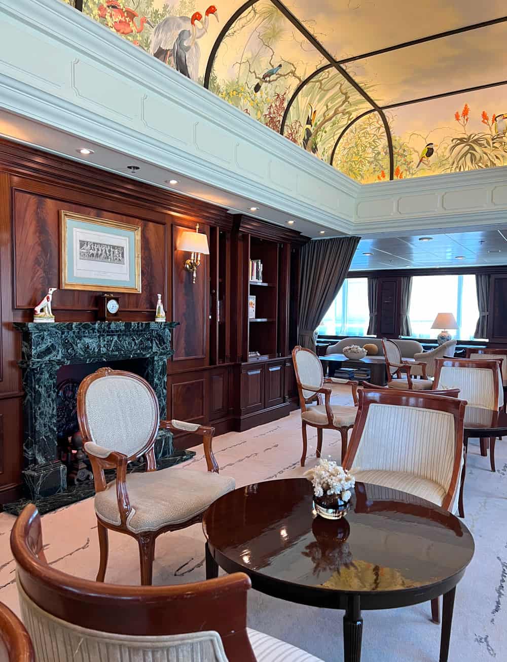 The Drawing Room lounge on Azamara Quest features dark wood paneling, a decorative marble fireplace, elegant armchairs and tables, and a painted ceiling mural with birds and tropical plants, creating a classic country-club atmosphere.