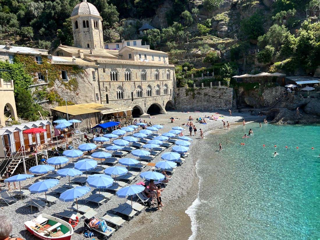 San Fruttuoso Beach on the Italian Riviera, with rows of blue beach umbrellas, crystal-clear turquoise water, and the historic Abbey of San Fruttuoso nestled against a wooded hillside.