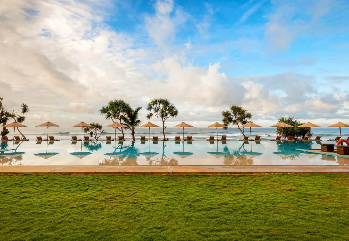 Green lawn in front of a turquoise infinity pool lined with loungers and umbrellas, overlooking the blue Indian Ocean