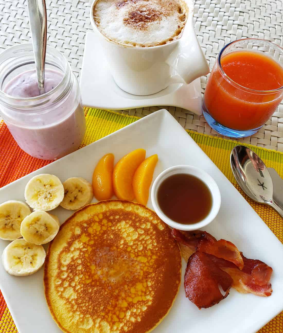 Pancakes with bacon, sliced banana and peaches, plus a glass of orange juice and a jar of pale purple blueberry yogurt