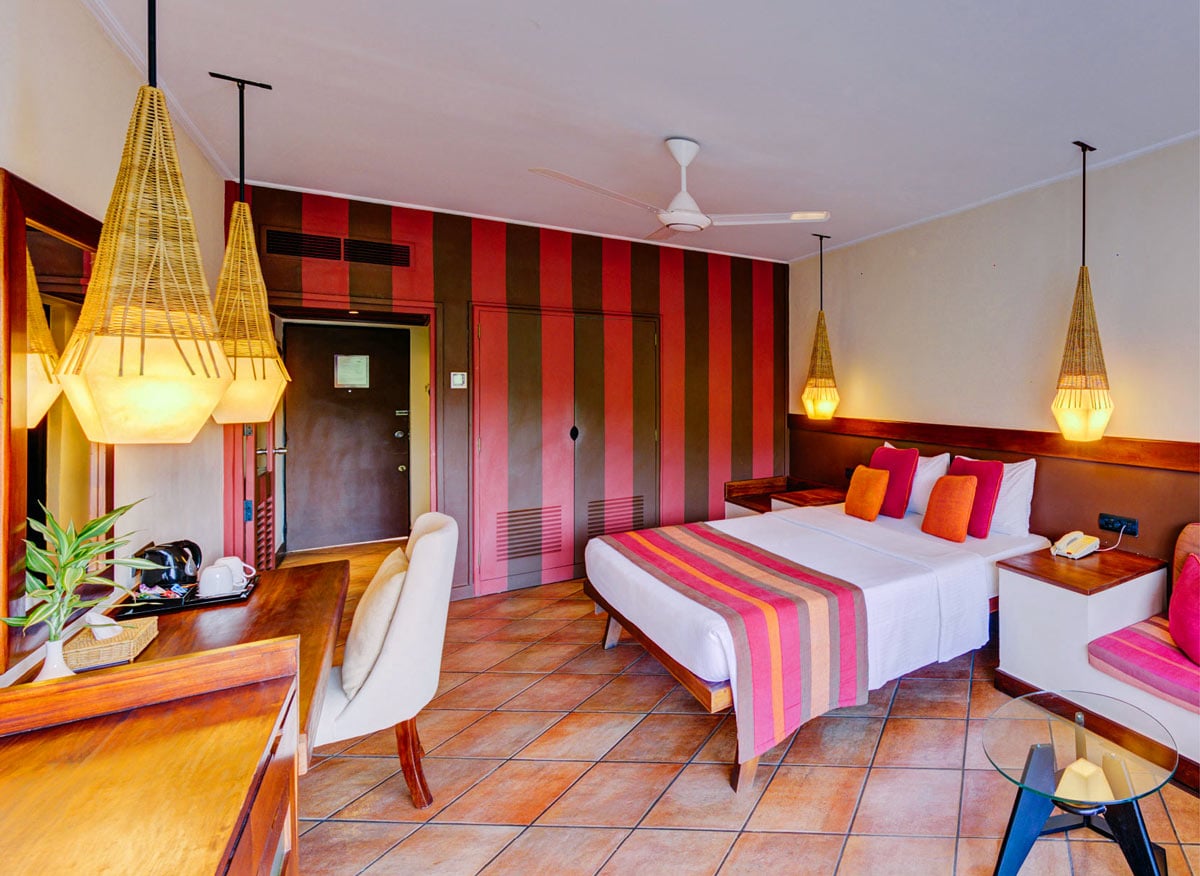 Guest room at Cinnamon Citadel Kandy with warm wood furnishings, terracotta-tiled floors, striped red accent wall and woven pendant lights
