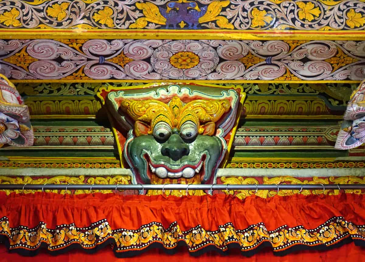 A colorful carved face with bulging eyes and white teeth, surrounded by intricate gold, green, red and purple painted patterns, inside the Temple of the Tooth in Kandy