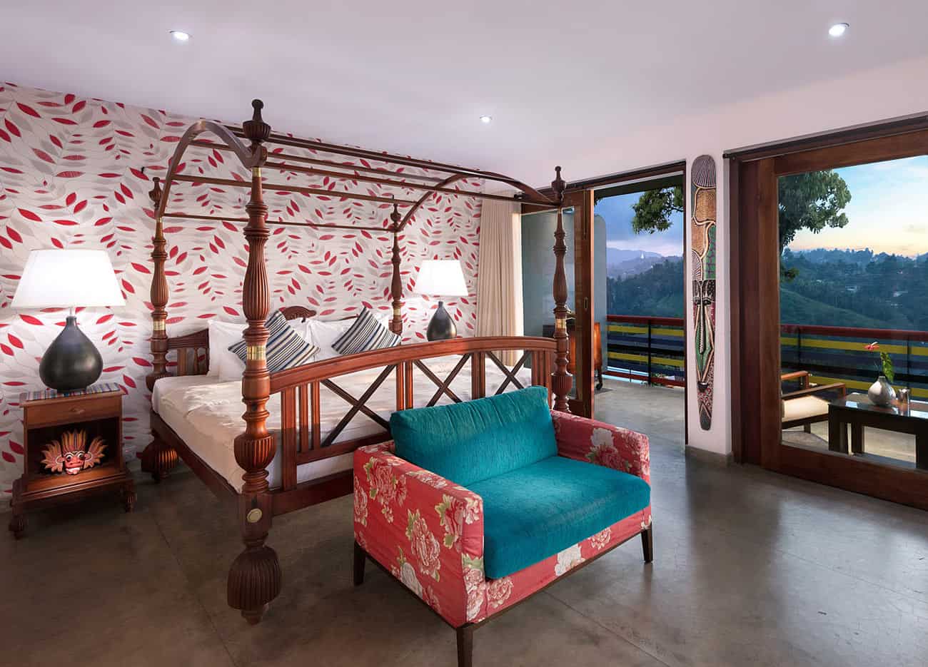 Pepper Deluxe Room at The Elephant Stables in Kandy, with a carved four-poster bed, red flower-covered chair and a private balcony overlooking green hills
