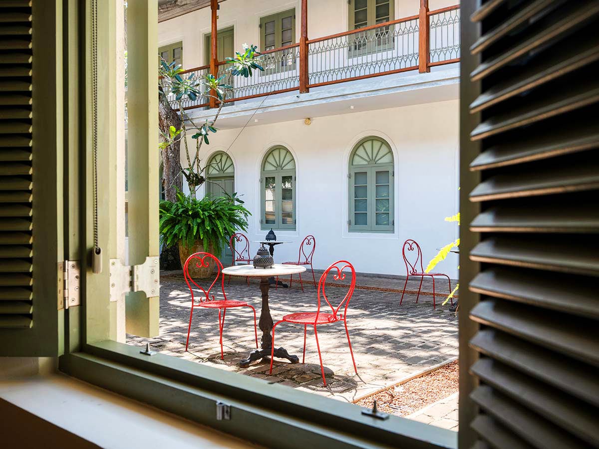 Red metal chairs and cafe tables in a courtyard by a white hotel building with green arched windows