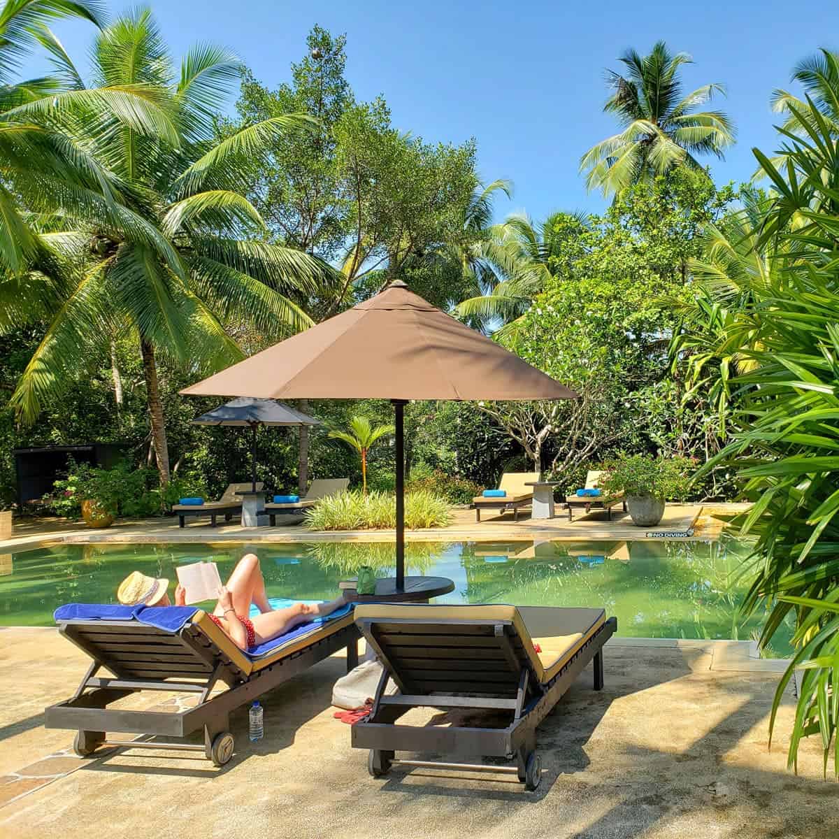 A woman in a red bikini reads on a shaded lounger beside a tropical garden pool.
