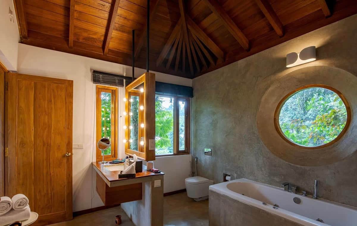 Modern bathroom with polished stone walls, a Jacuzzi tub, warm wood ceiling and round porthole window looking out to greenery