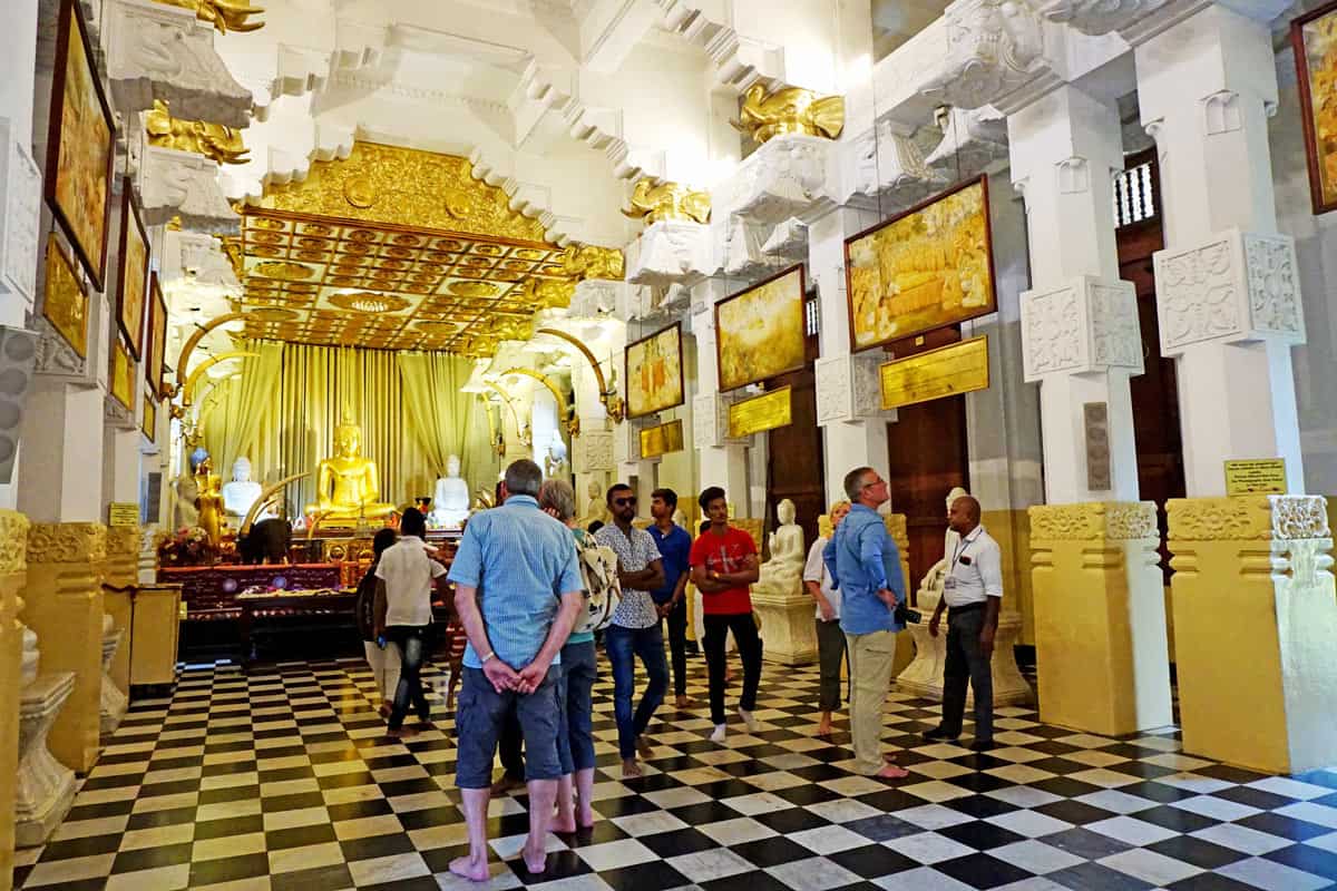 Visitors throng around in the "New Palace" hall, with its black-and-white tiled floor, white carved columns, golden ceiling decorations, framed paintings and a glowing gold Buddha statue at the far end.