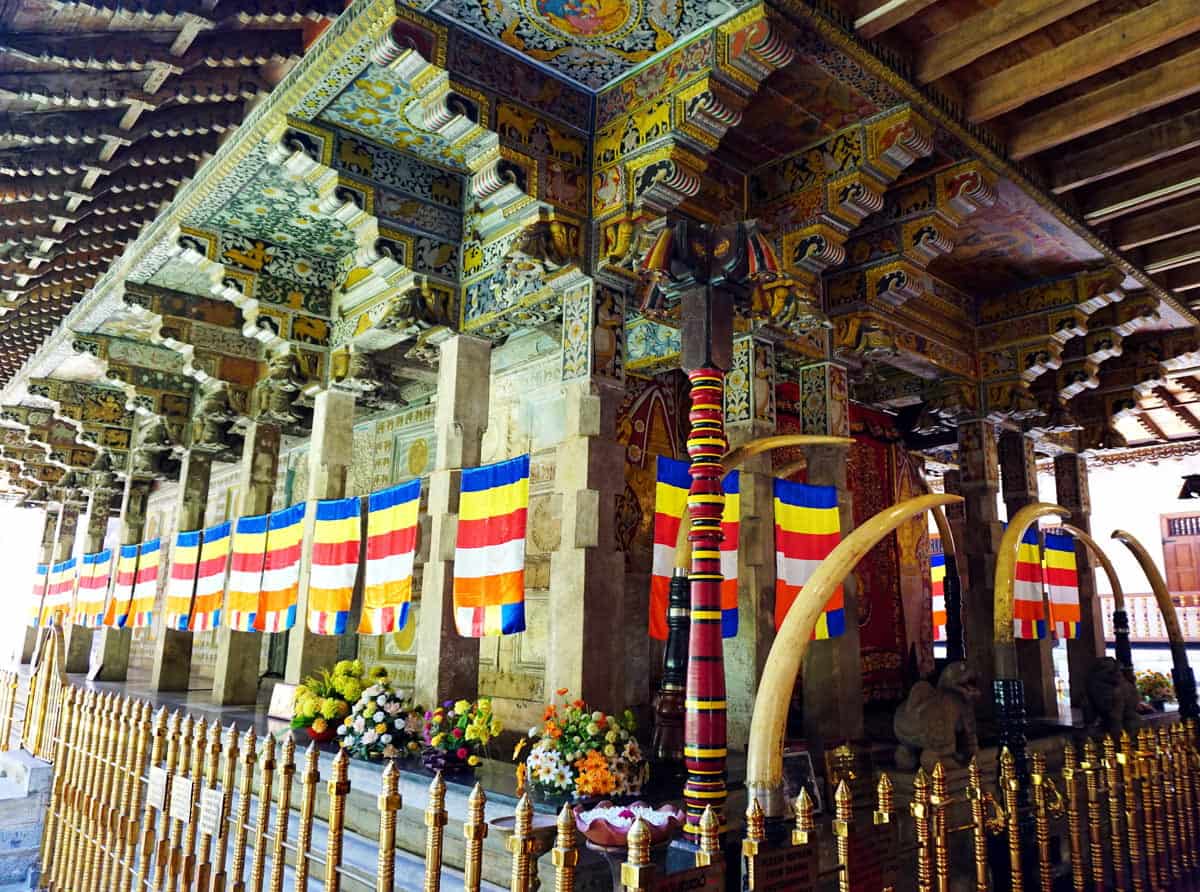 Ornately decorated Buddhist shrine with colorful flags, flower offerings, golden railings and four enormous curved elephant tusks flanking the sacred relic chamber.