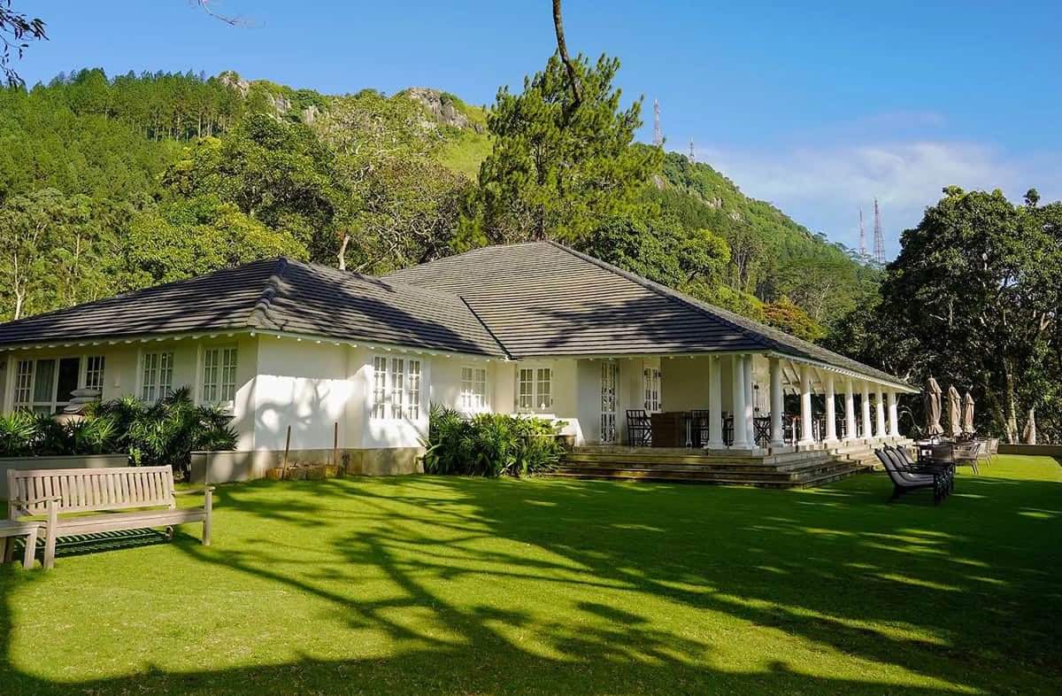 White colonial-style W15 Hanthana Estate bungalow with a pillared veranda, manicured lawn and forested hills behind