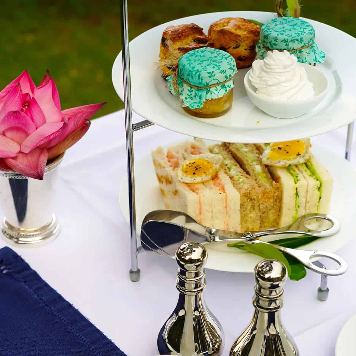 Afternoon tea tower with savory sandwiches, scones, jam and cream and a pink flower on the side