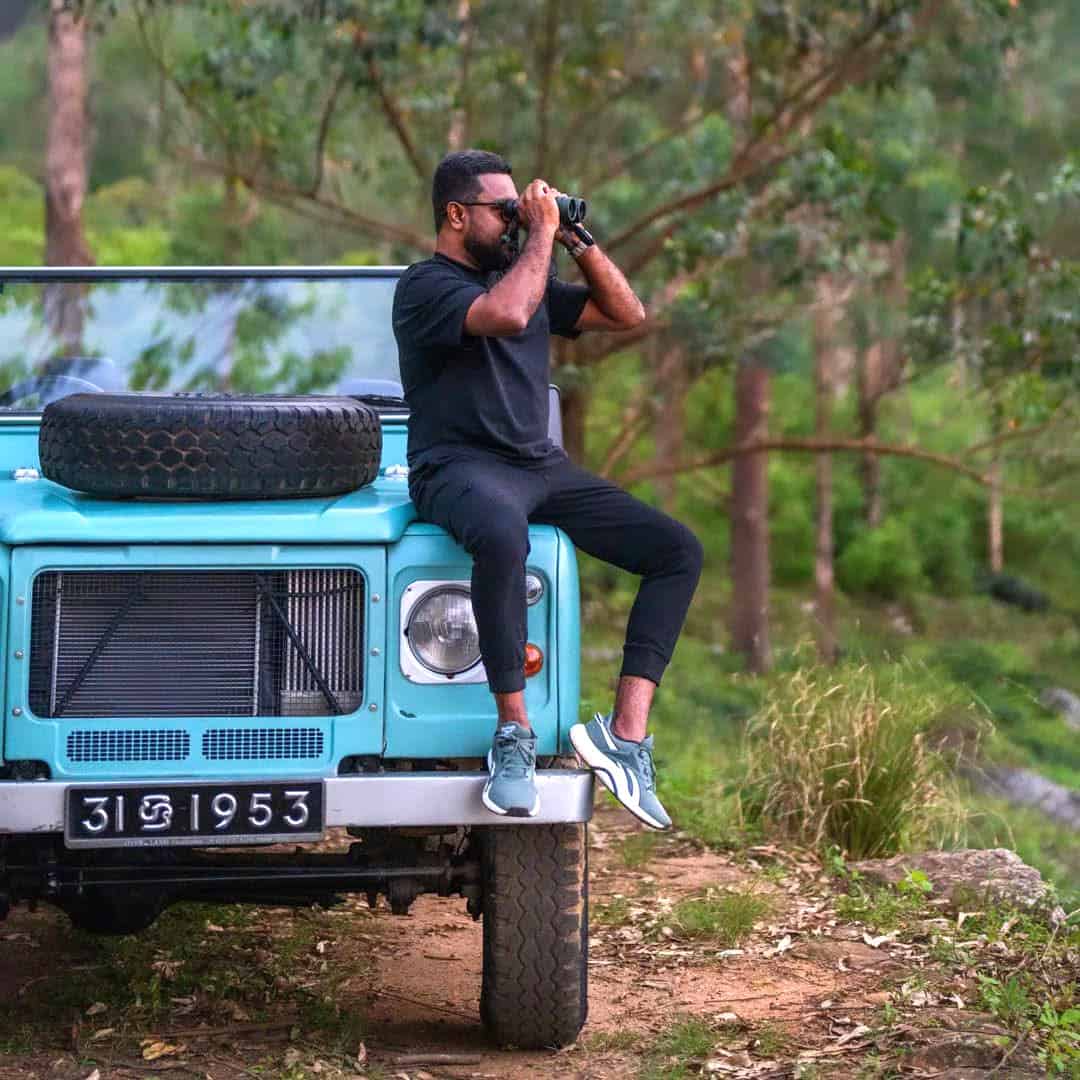 Naturalist looks through binoculars, while sitting on the hood of a vintage turquoise Land Rover in the forested Hanthana Hills.