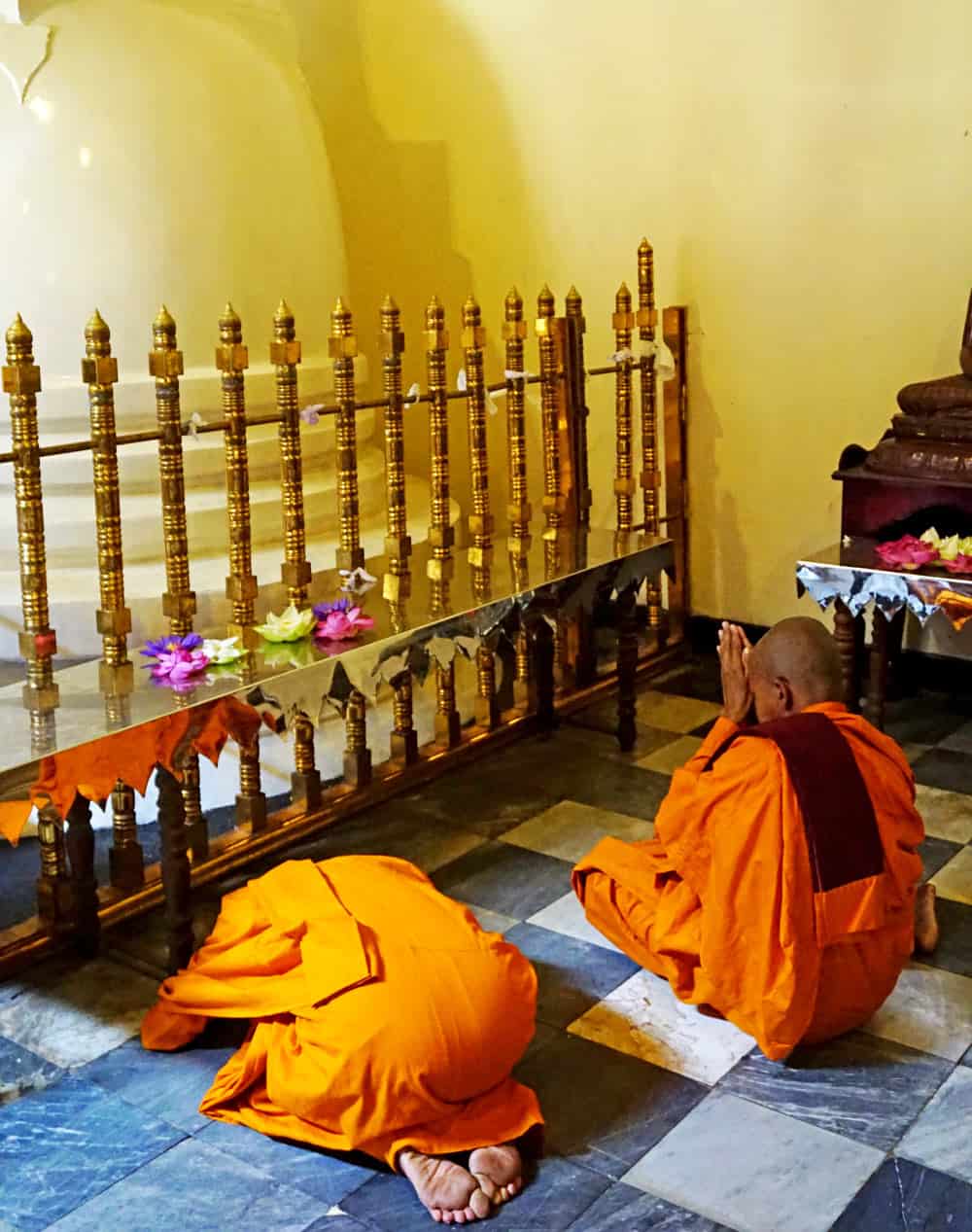 Two Buddhist monks in orange robes kneel and bow in prayer on a black-and-white tiled floor beside a gleaming gold railing scattered with lotus flowers. 