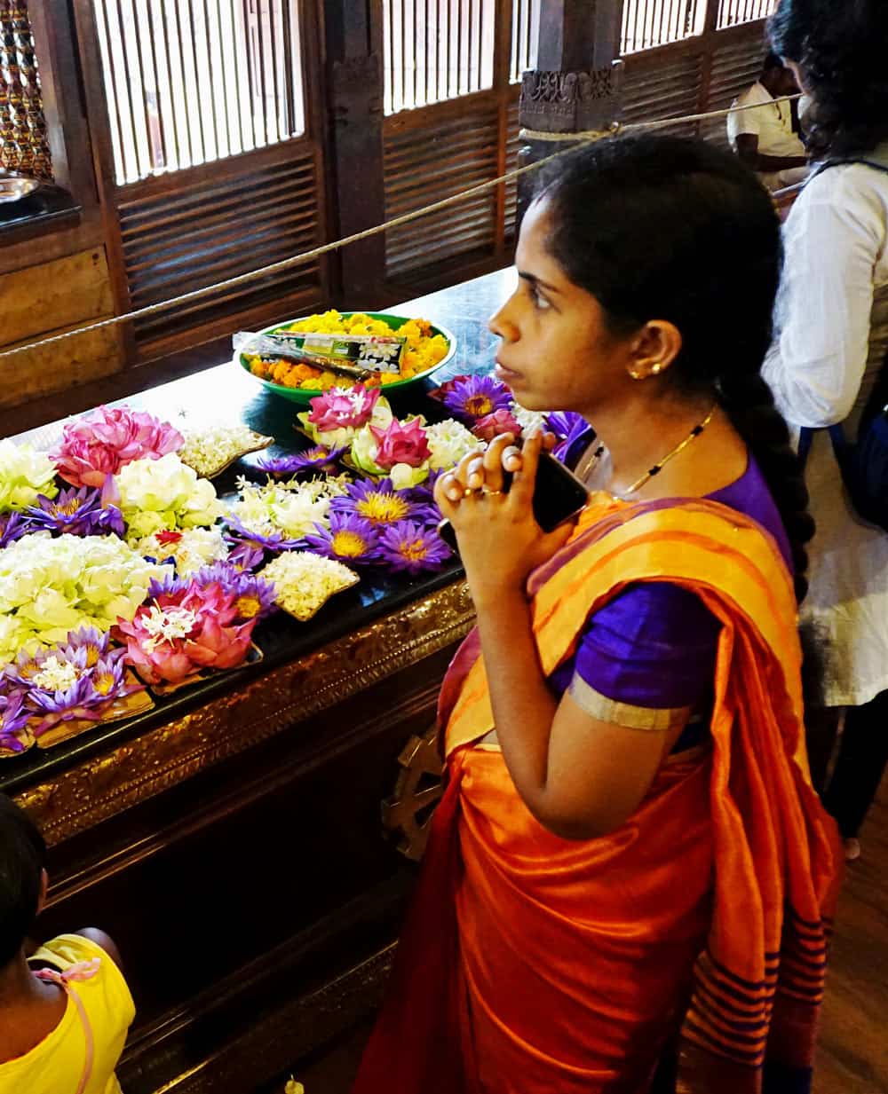 A young woman in an orange sari stands beside a Buddhist temple table overflowing with pink, purple, yellow and white lotus flowers.